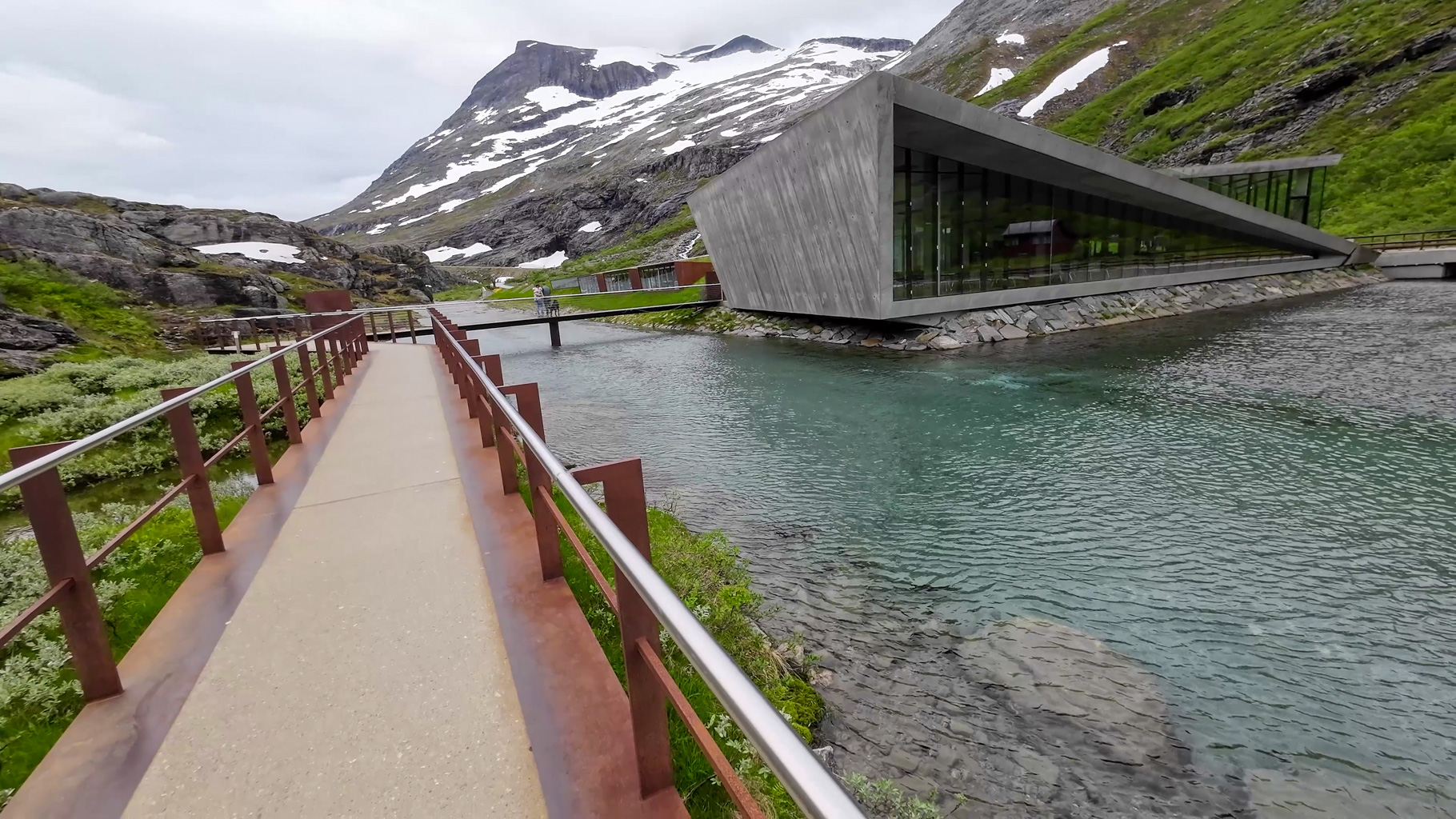 Betónová budova s ostrými líniami a odrazom hôr v oknách. Budova turistického centra Trollstigen pri rieke v horskom prostredí.