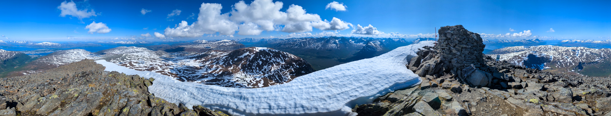 Panoramatický záber z vrcholu odhaľuje 360-stupňový výhľad na okolité fjordy, horské chrbty a zasnežené štíty. Vpravo vidieť aj vrcholový bod označený mohylou.