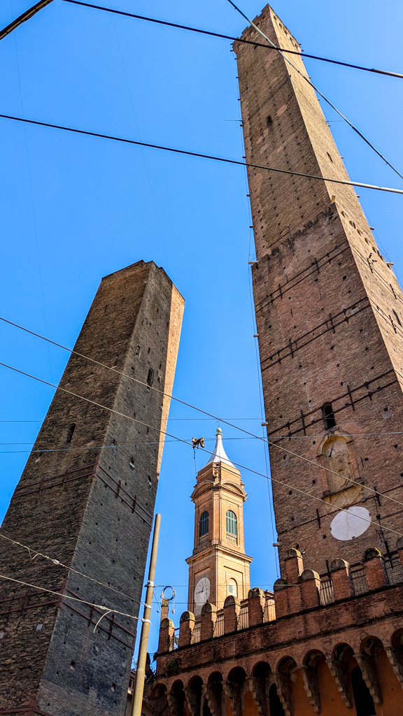 Pohľad na ikonické Šikmé veže v Bologni – Torre degli Asinelli a Torre Garisenda. Tieto stredoveké veže boli symbolom prestíže bohatých rodín a slúžili aj ako obranné stavby. Torre Garisenda je známa svojim výrazným náklonom, ktorý je ešte väčší než v Pise.
