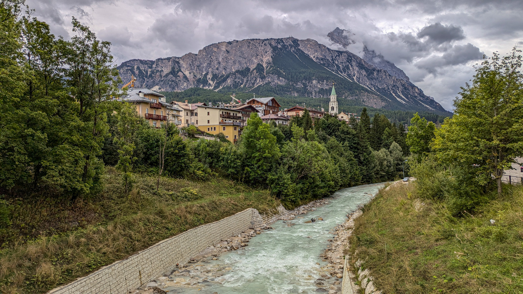 Malebný pohľad na Cortinu d'Ampezzo s prúdiacou riekou Boite, obklopenú mohutnými štítmi Dolomitov. V popredí vyniká kostol s typickou zelenou vežou.