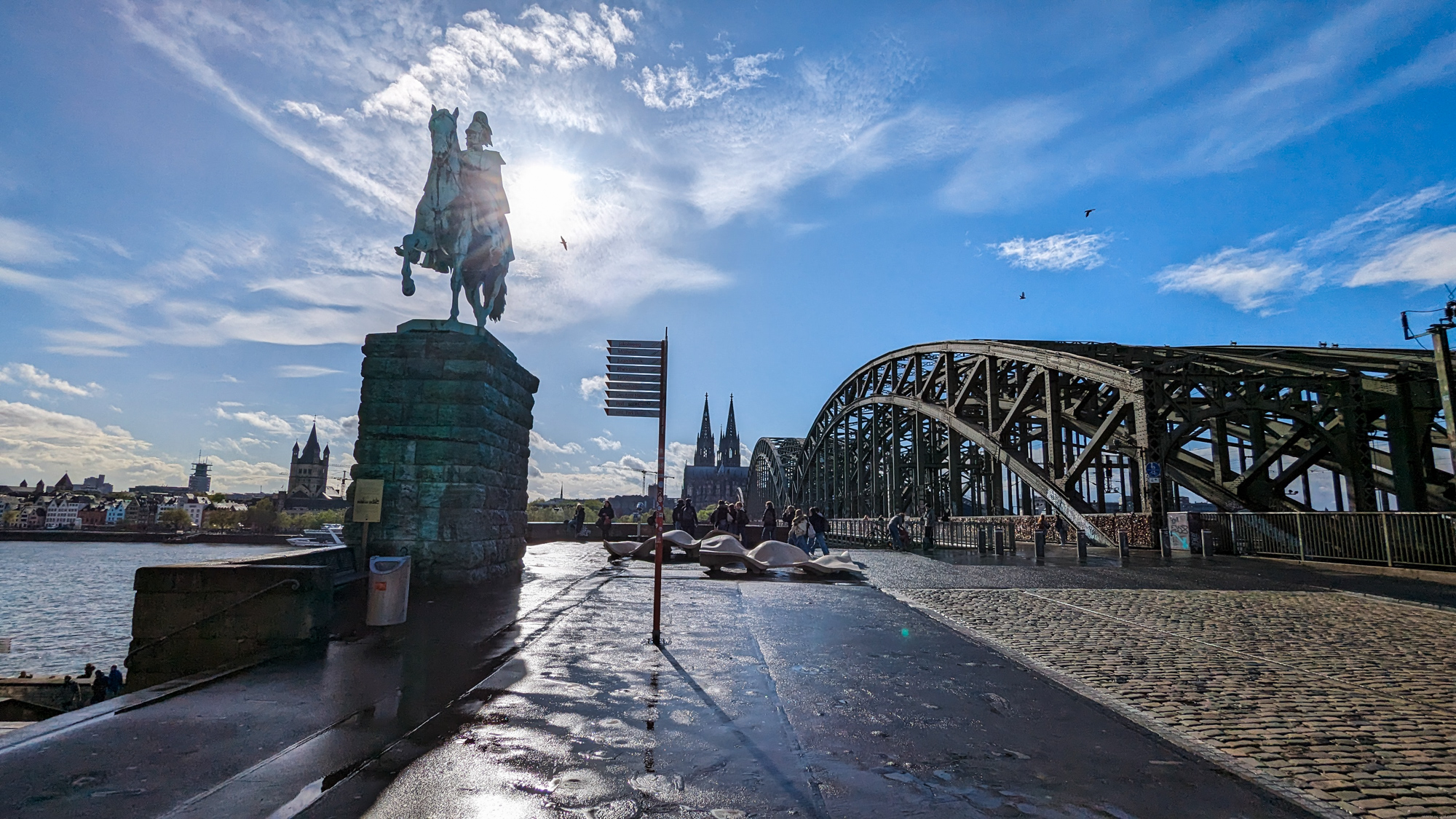 Pohľad z mosta na jazdeckú sochu a most Hohenzollernbrücke, s katedrálou v diaľke proti ostrému slnku.